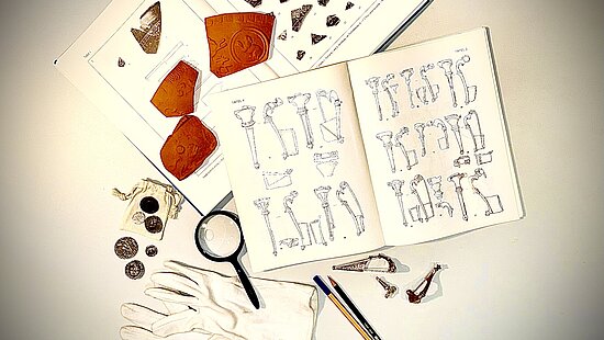 Fibeln, Münzen und Sigillatascherben mit Lupe, Hanschuhen und Fachliteratur